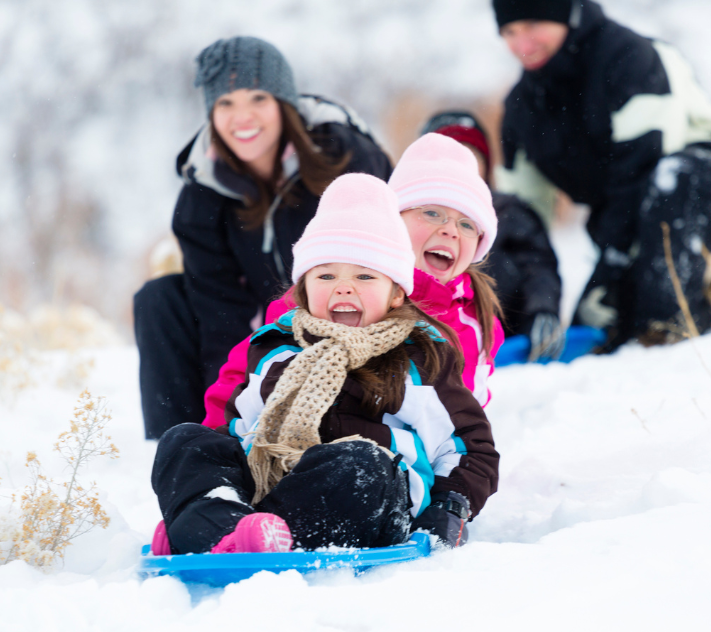 Two young girls sledding down a hill with their mother in the background