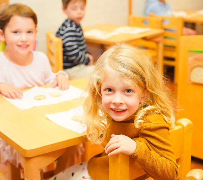 young girl smiling while sitting at a desk; other children in background