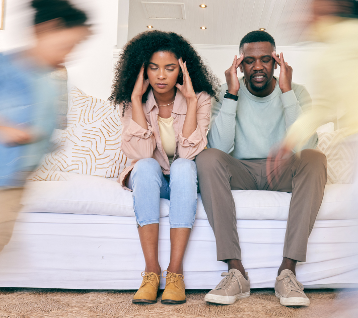 Two parents sitting on the couch holding their heads while their children are running around them