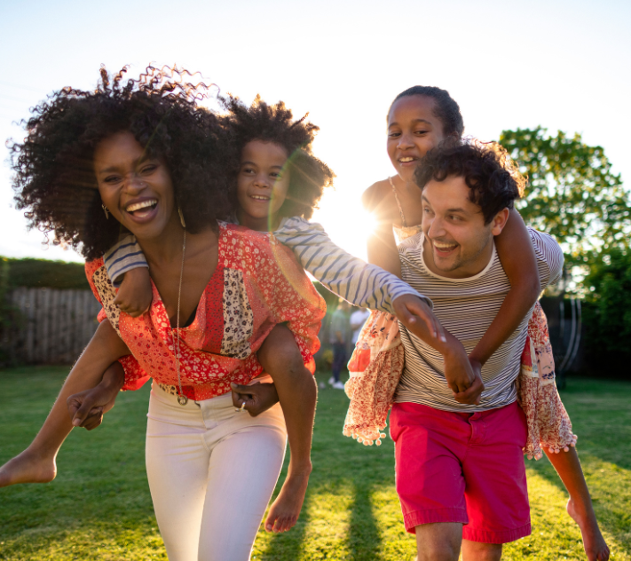 Mother with daughter on her back and father with daughter on his back laughing and smiling together in a backyard.