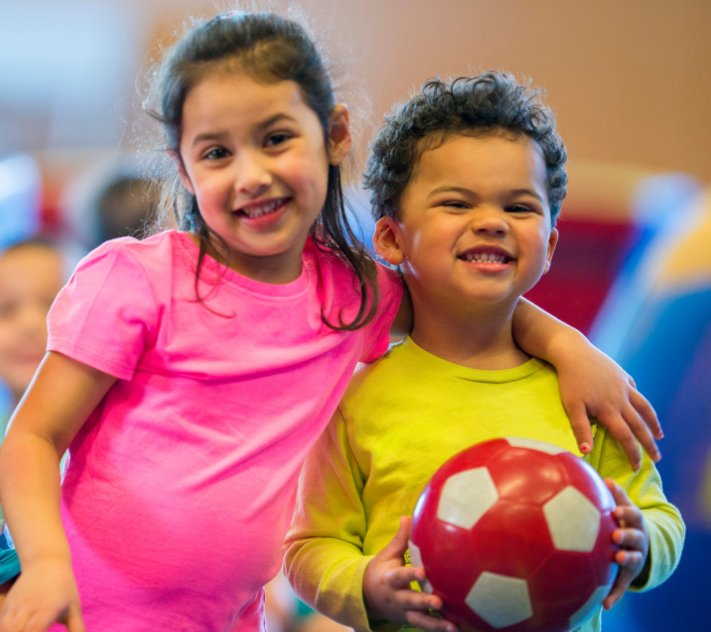 Young girl with her arm around a shorter young boy who is holding a red soccer ball