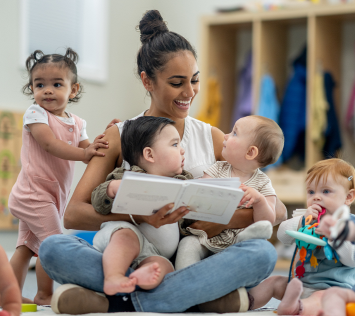 Teacher sitting on the floor with a holding a toddler and a book with other toddlers around her.